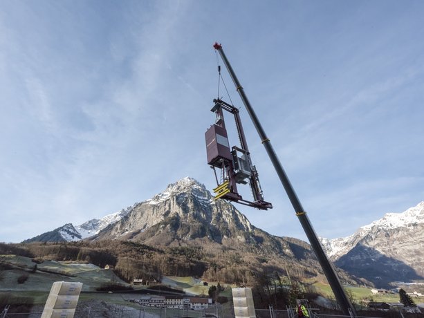 Ein Regalbediengerät schwebt an einem Kran über einem Hochregallager in der Schweiz, im Hintergrund Berge